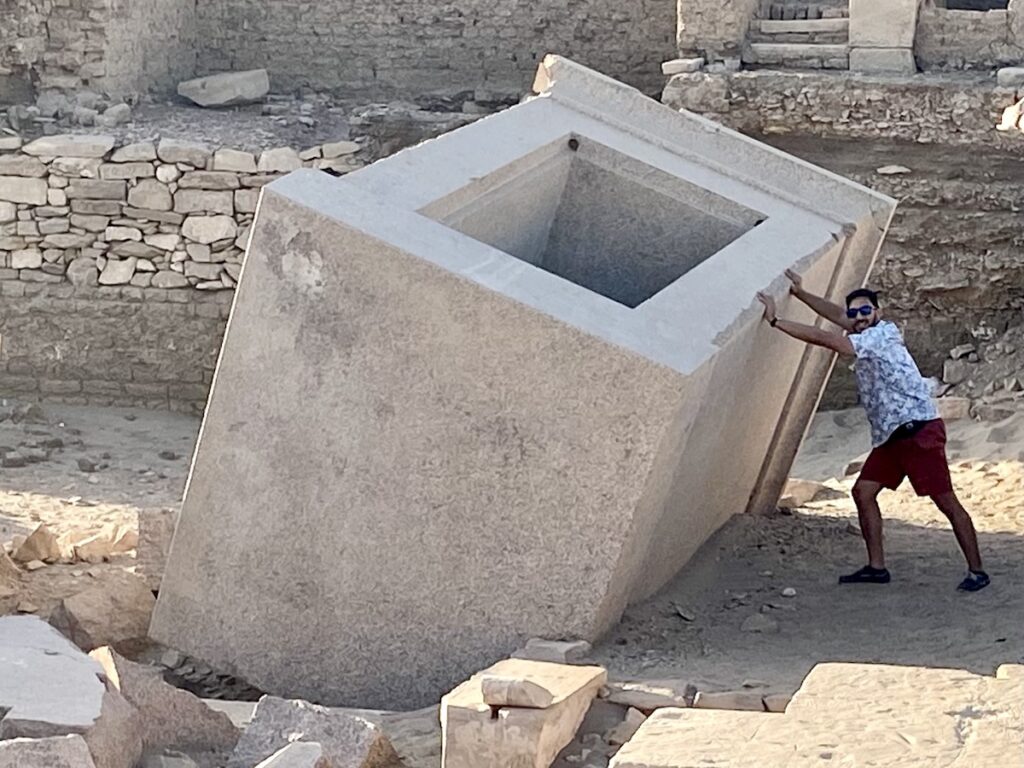 Large tilted granite naos on Elephantine Island in Aswan, Egypt, with a visitor standing beside it for scale, showing the hollow rectangular interior of the ancient temple shrine.