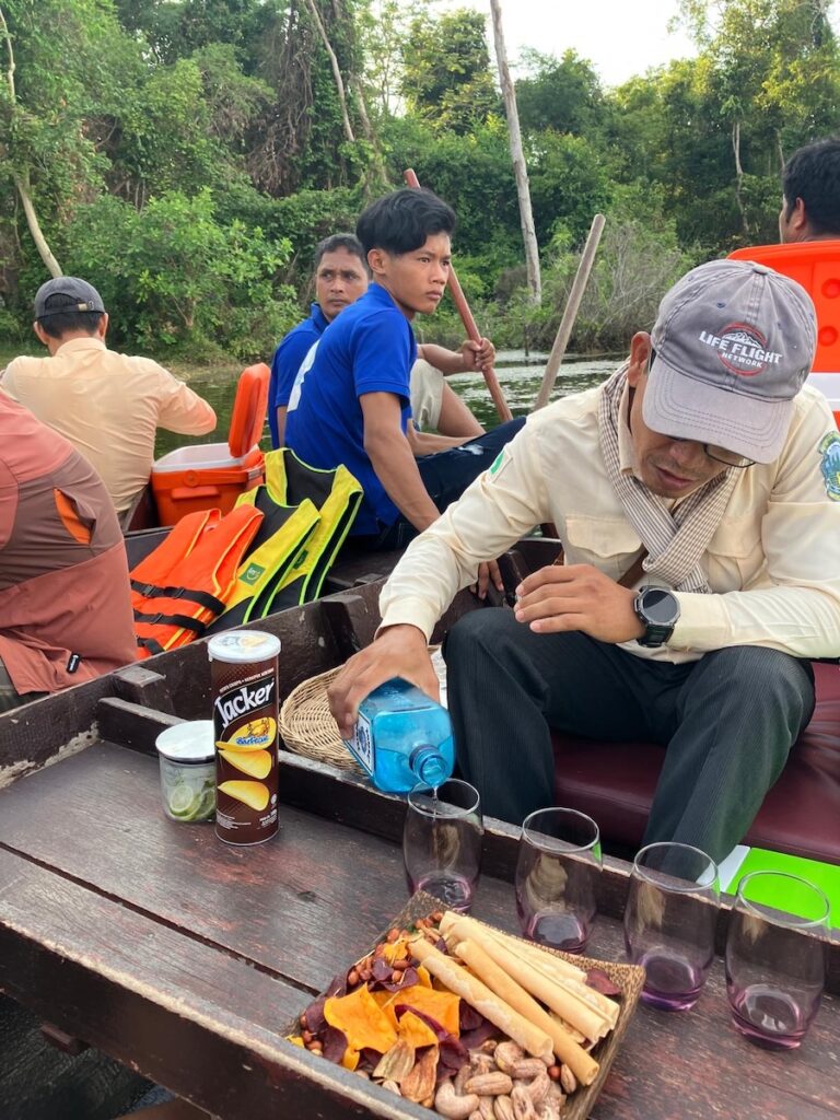 A group of people enjoying a sunset boat ride on the Angkor Thom moat in Siem Reap, Cambodia. A guide pours drinks beside a wooden tray with glasses, snacks, and chips, as others paddle and relax amid lush greenery.