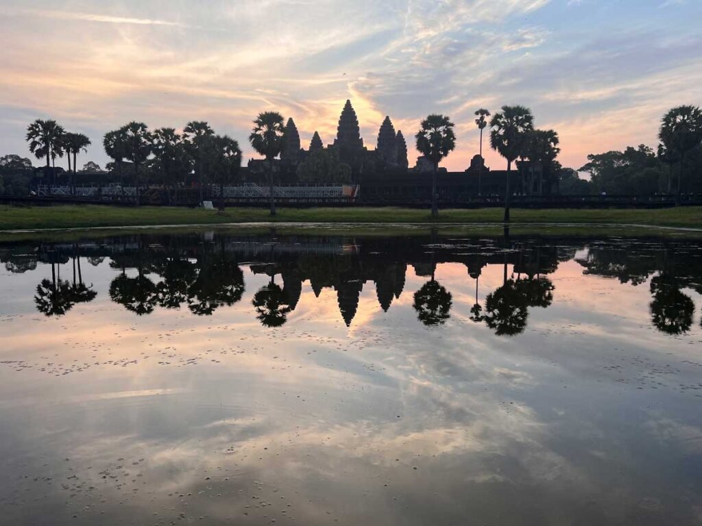 Angkor Wat silhouetted at sunrise with vibrant skies and palm trees reflecting in the still waters of the lotus pond, captured from the iconic viewpoint in Siem Reap, Cambodia.