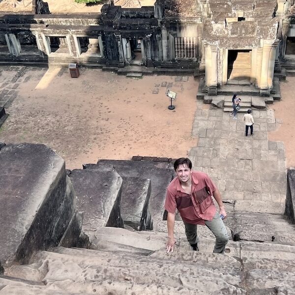 Luke Caverns climbs a steep stone staircase at Ta Keo, surrounded by ancient temple architecture and weathered carvings in the background.