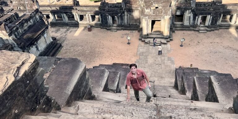 Luke Caverns climbs a steep stone staircase at Ta Keo, surrounded by ancient temple architecture and weathered carvings in the background.