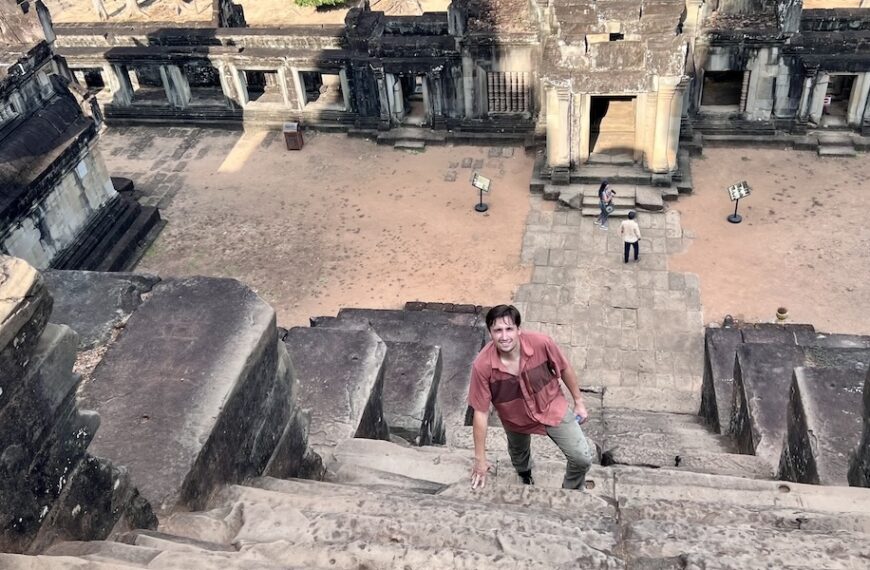Luke Caverns climbs a steep stone staircase at Ta Keo, surrounded by ancient temple architecture and weathered carvings in the background.