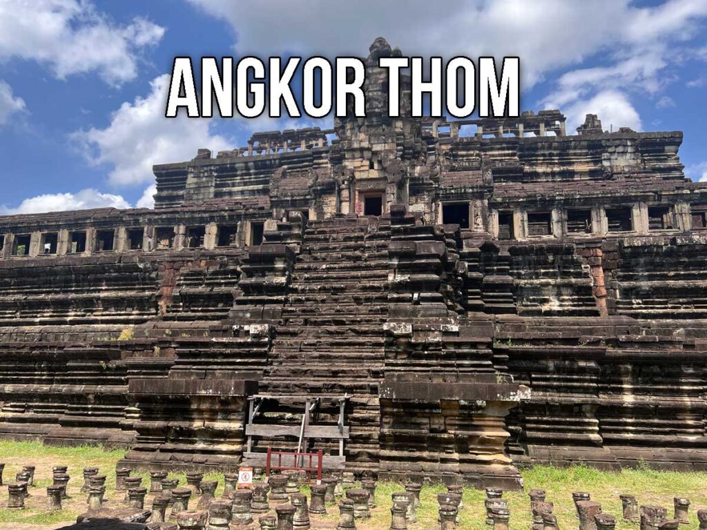 Front view of a large stepped temple inside Angkor Thom, showing dark stone terraces, aligned columns, and a stairway rising to the upper level under scattered clouds and blue sky.