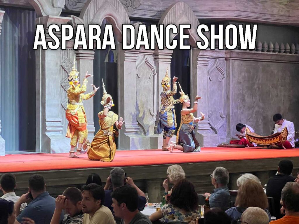 Cambodian dancers in ornate costumes perform a classical Apsara dance on stage, with musicians playing traditional instruments and a seated audience watching closely.