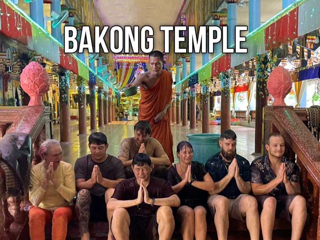 A group of visitors sits respectfully with hands pressed together after receiving a blessing from a Buddhist monk inside a vibrant hall near Bakong Temple, surrounded by colorful decorations and temple columns.