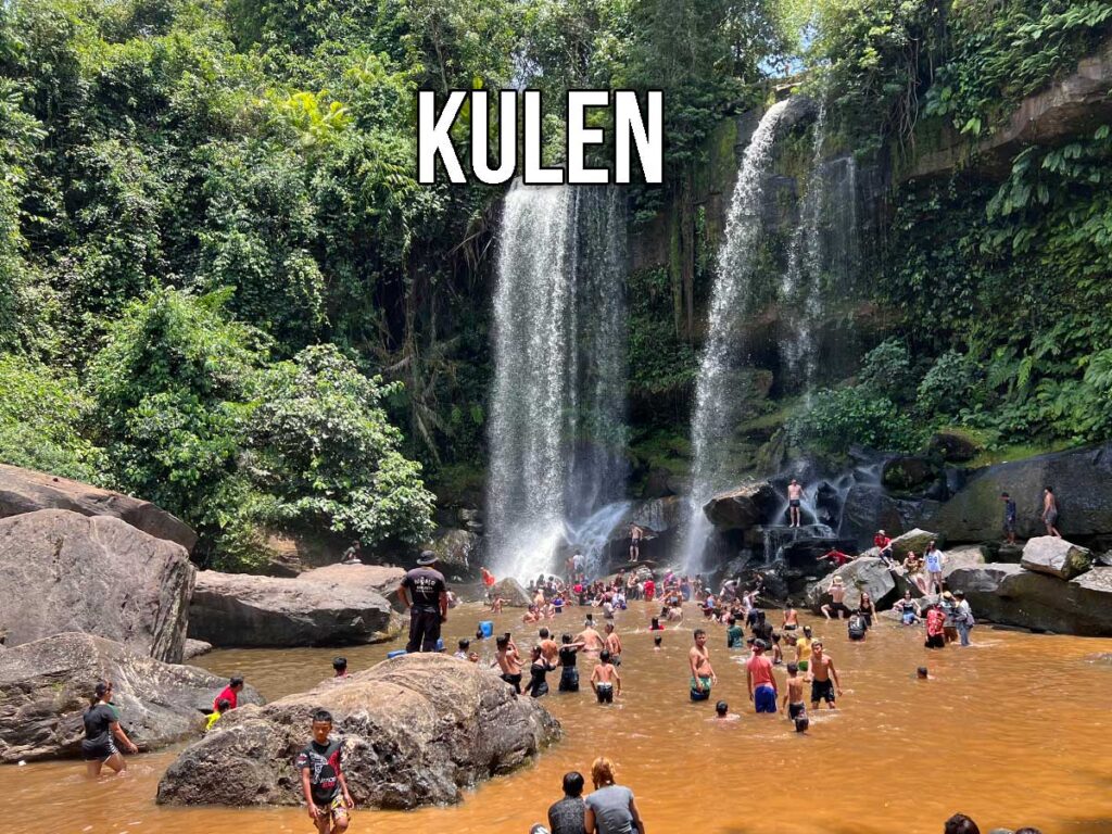 Large crowd swimming and relaxing in the reddish pool beneath the twin waterfalls of Kulen Mountain, surrounded by dense jungle and towering rock walls.