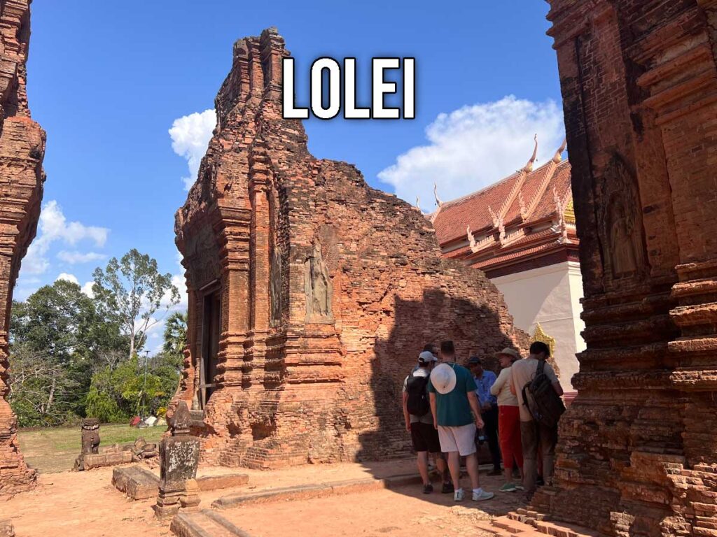 Tour group standing near the red brick towers of Lolei Temple, with a traditional Buddhist monastery visible behind the ancient Khmer ruins under a bright blue sky.