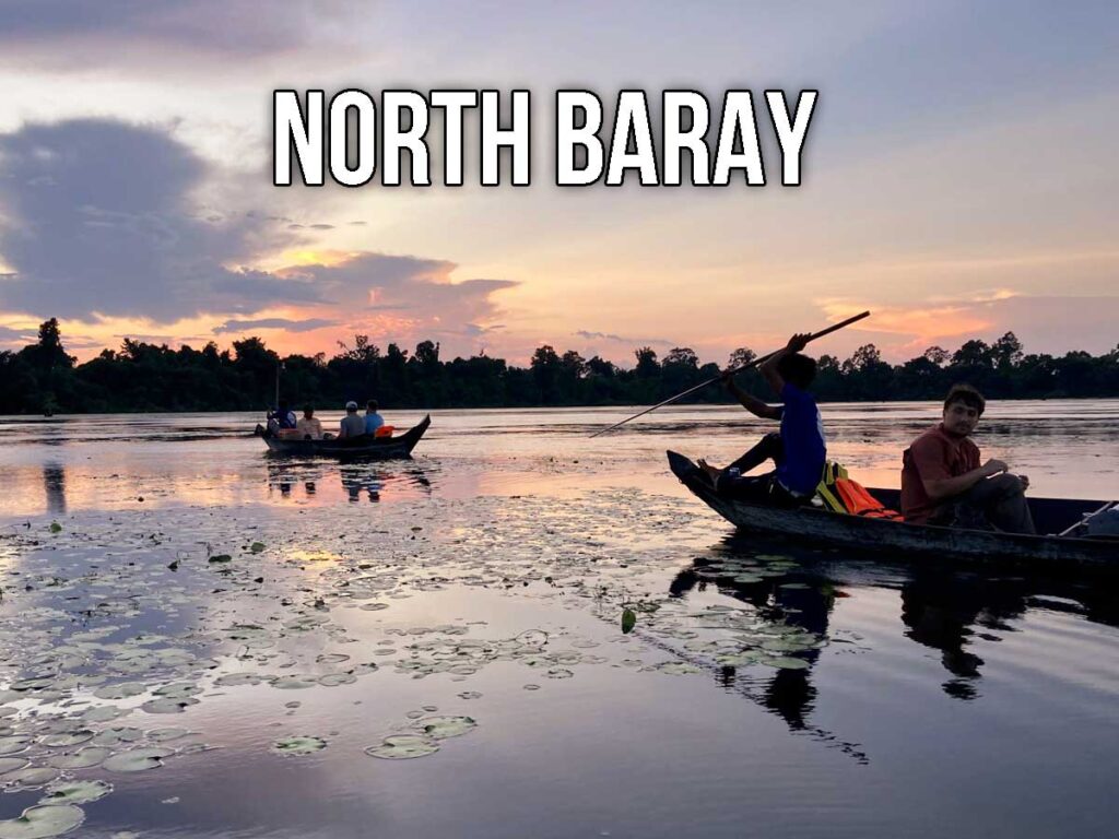 A boatman guides a small wooden canoe across the lotus-dotted moat of Angkor Thom at sunset, with visitors relaxing on board and the sky glowing in soft shades of orange and blue.