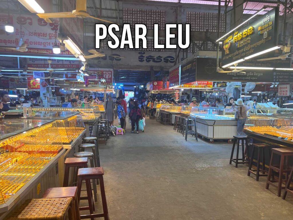 Interior of Psar Leu Market with bright glass jewelry counters, vendors, and shoppers walking through narrow aisles under fluorescent lighting.