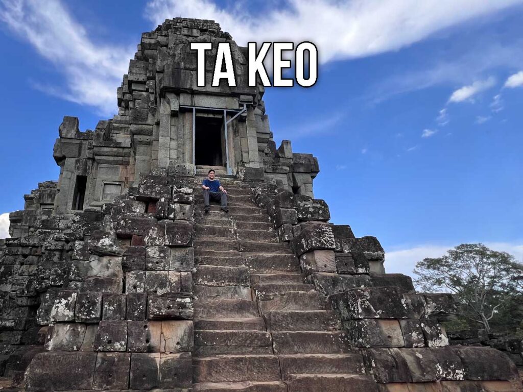 A visitor sits halfway up the steep central staircase of Ta Keo temple, surrounded by massive sandstone blocks under a clear blue sky.