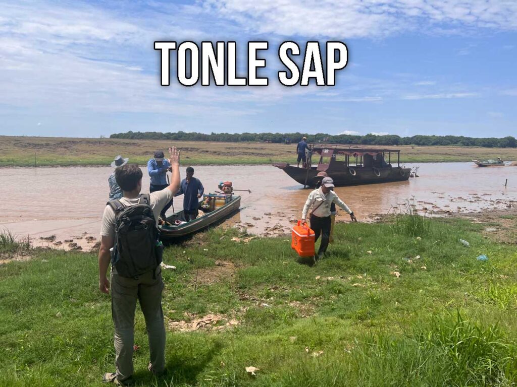 Tour participants gather on the grassy banks of Tonle Sap as boats wait in the shallow muddy water under a bright sky, with one guide carrying an orange cooler.