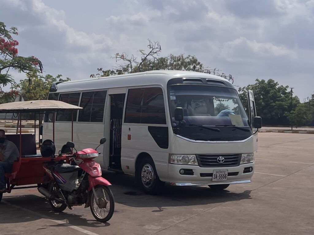 hite 13-seat Toyota Coaster minibus parked under trees near a roadside area in Cambodia, used for transporting participants on the Angkor Zenith Passage Tour.