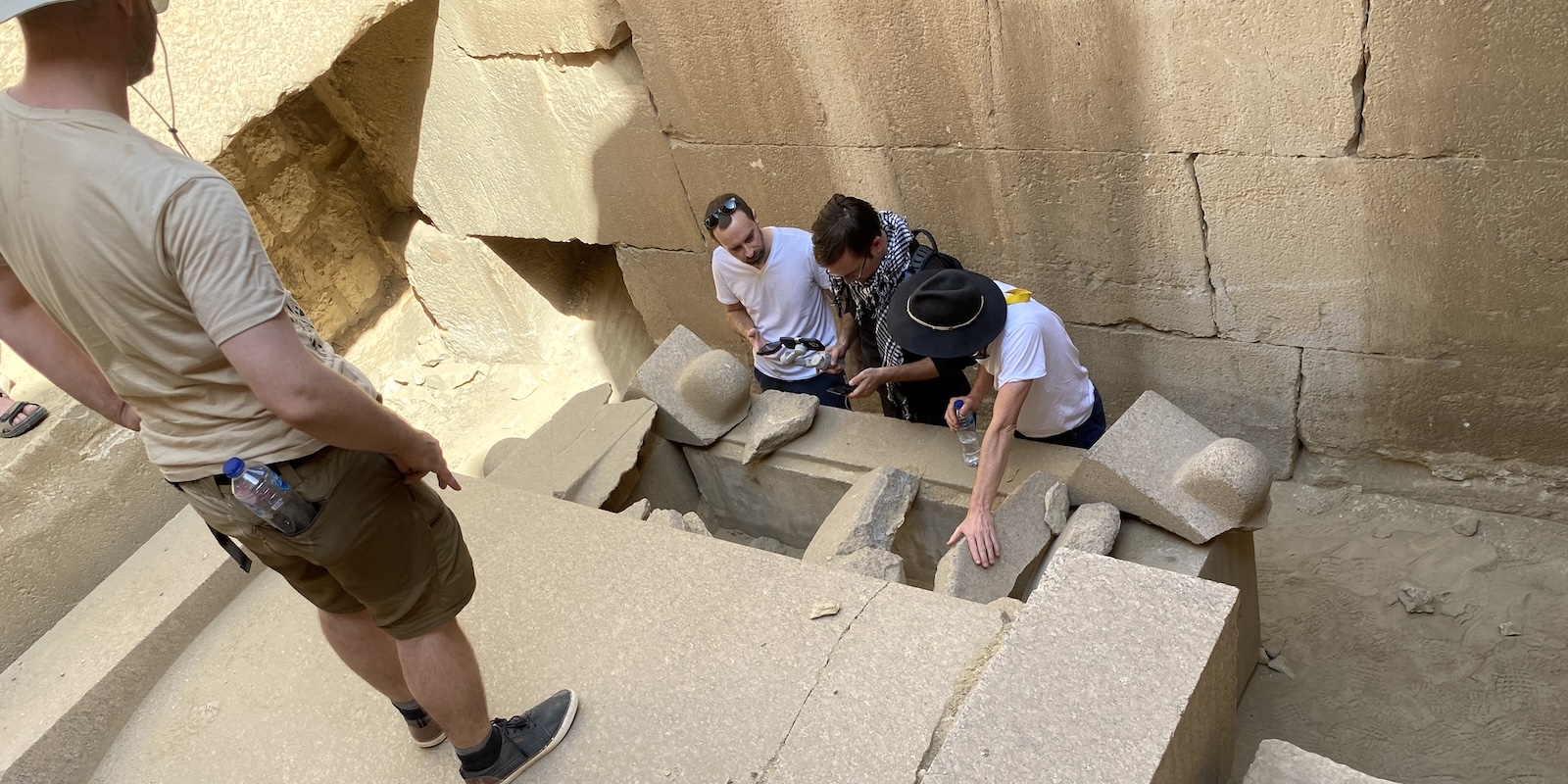 Broken stone sarcophagus at Abu Ghurab in Egypt, with visitors inspecting carved stone fragments near the Sun Temple of Niuserre in the Abusir-Saqqara area.
