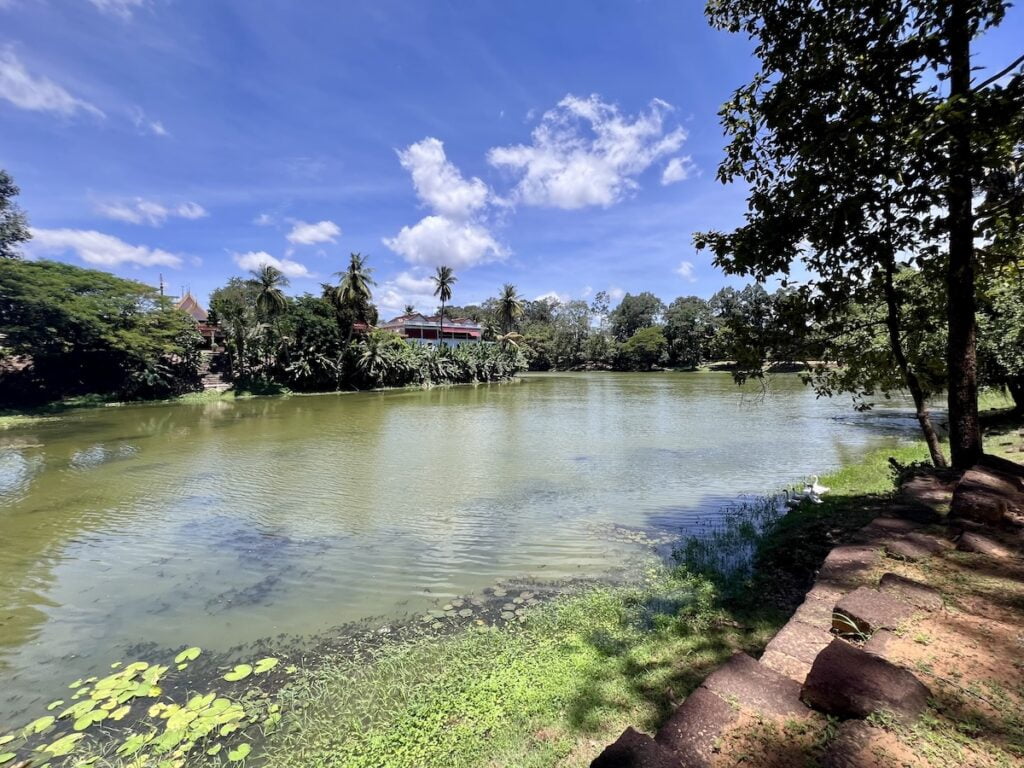 Wide view of the moat surrounding Bakong temple in Cambodia with tropical trees, still water, and the ancient Khmer temple complex beyond the shoreline