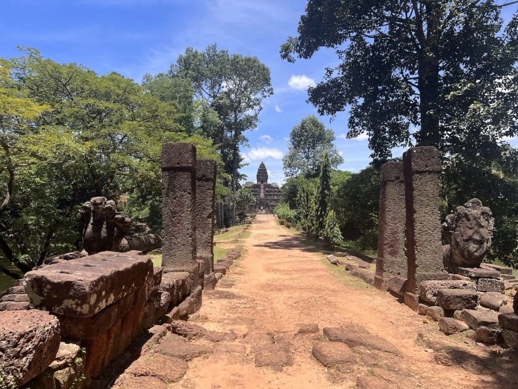 Ancient stone-lined causeway leading toward Bakong temple in Cambodia framed by guardian statues, laterite pillars, and dense tropical trees under a bright blue sky