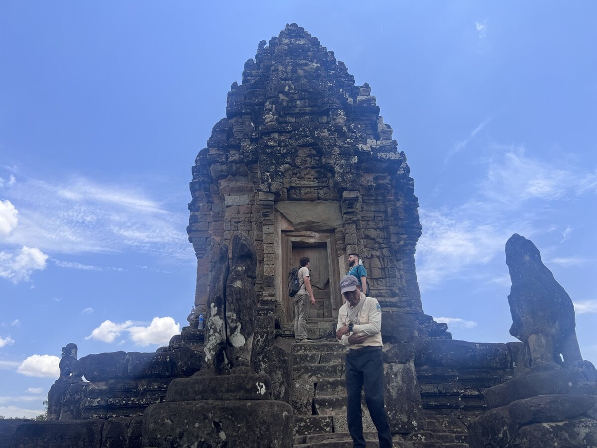 Visitors standing beside the central sanctuary tower at the summit of Bakong temple in Cambodia with steep stone steps, weathered carvings, and guardian statues beneath a bright blue sky