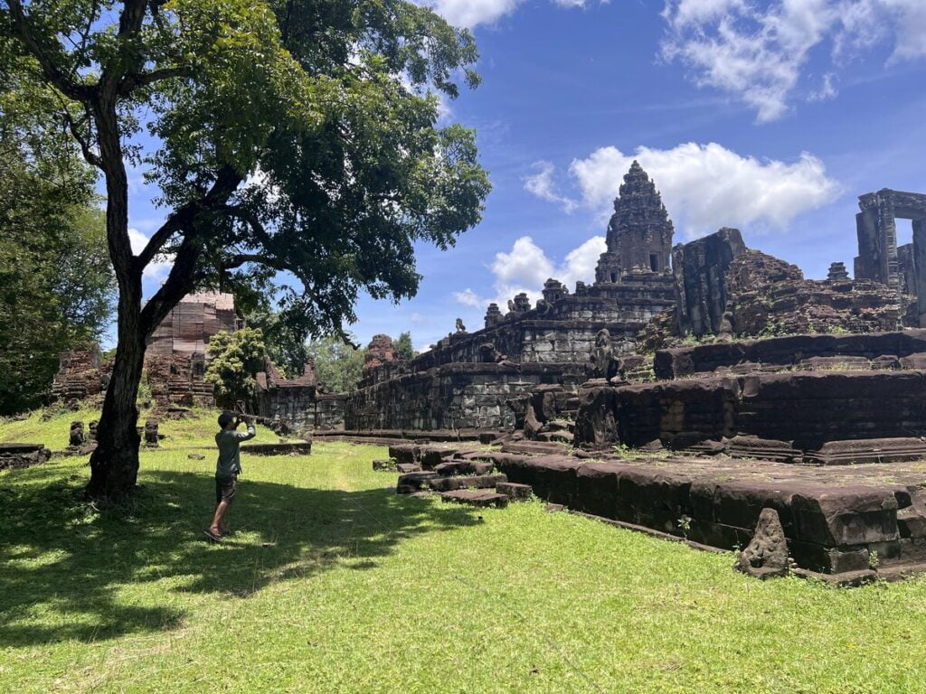 Wide exterior view of Bakong temple in Cambodia showing the stepped pyramid terraces, central sanctuary tower, grassy enclosure, and surrounding trees under midday sunlight