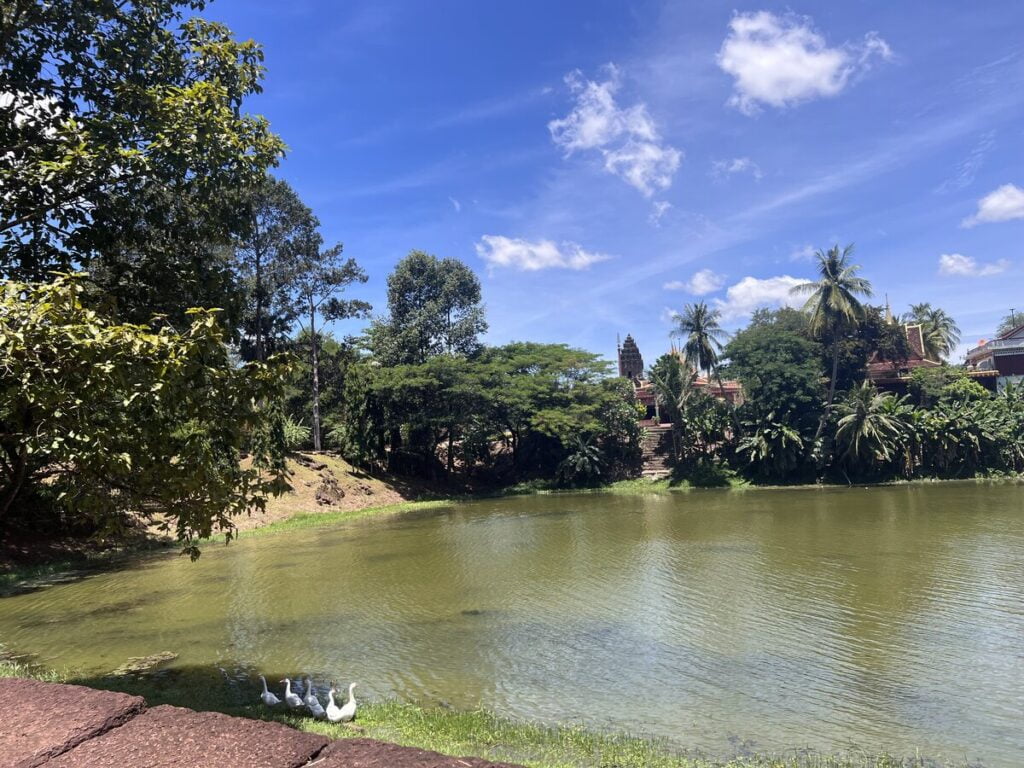 View across the moat surrounding Bakong temple in Cambodia with tropical trees, calm water, and the ancient Khmer temple rising in the background