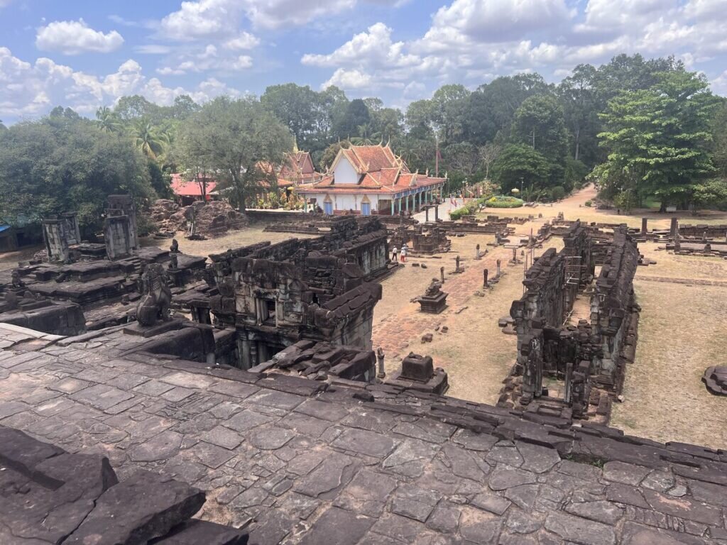 Elevated view from Bakong temple in Cambodia overlooking ancient stone enclosures, ruined galleries, and a modern Buddhist monastery beside the historic Khmer complex