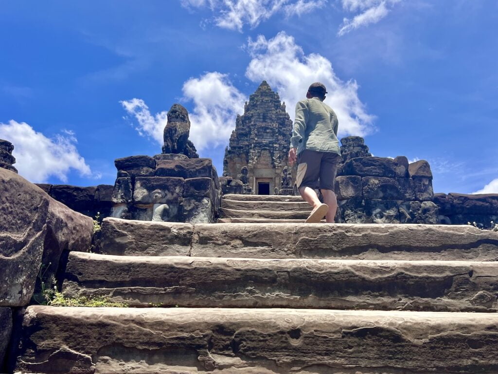 Visitor ascending the steep stone steps toward the central sanctuary tower of Bakong temple in Cambodia beneath a bright blue tropical sky