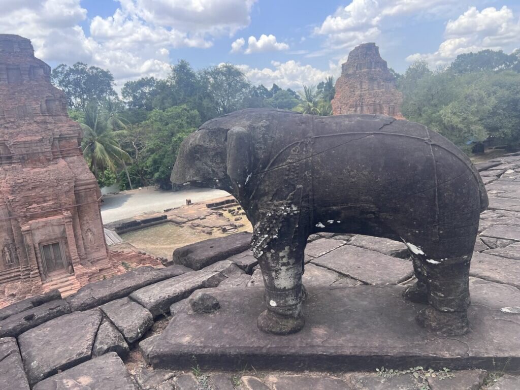 Ancient stone elephant statue on the upper terrace of Bakong temple in Cambodia overlooking brick towers and the surrounding Khmer temple complex under a cloudy sky