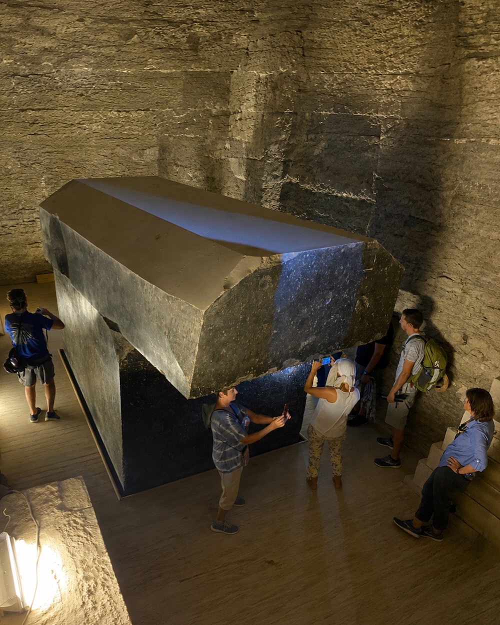 Visitors standing beside one of the enormous polished granite sarcophagi inside the underground chambers of the Serapeum of Saqqara in Egypt showing the massive scale and precision stonework of the ancient structure