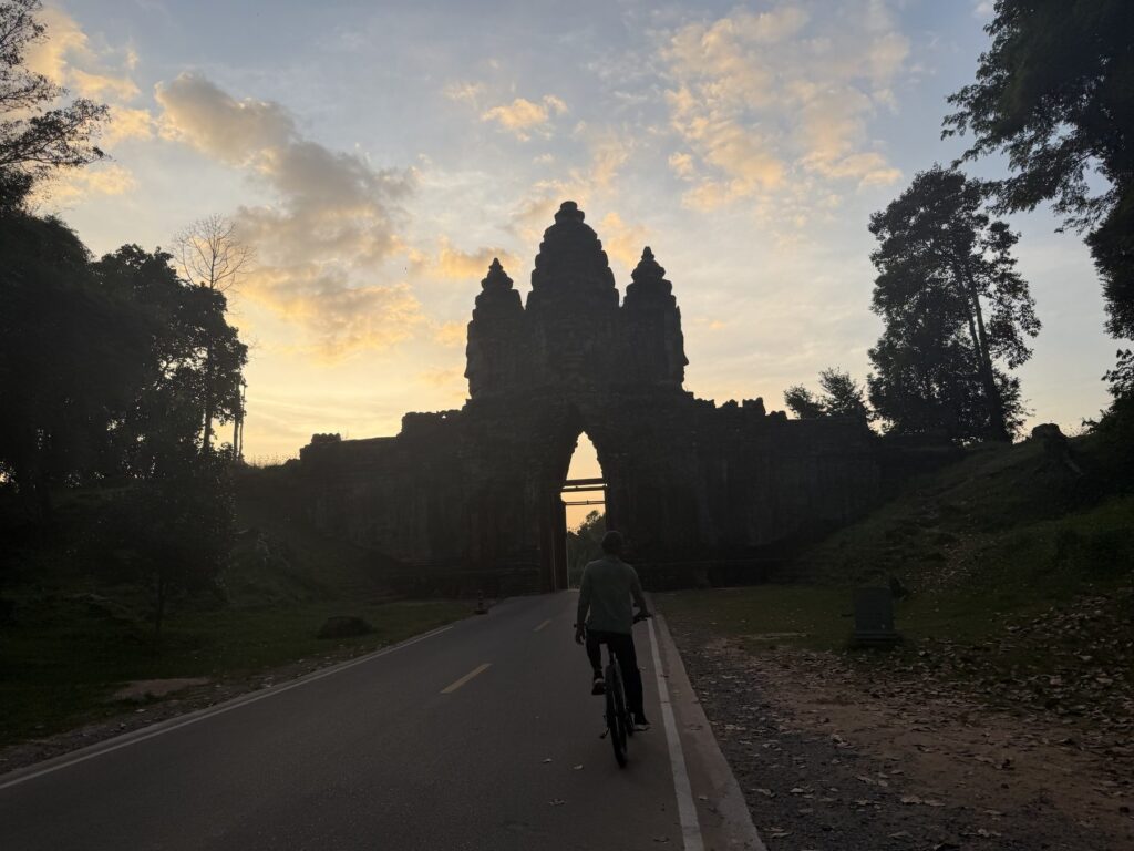 Guide Kanha Phum cycling toward the silhouetted West Gate of Angkor Thom at sunset