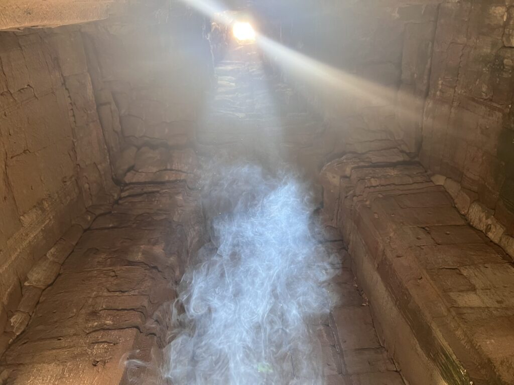 Incense smoke drifting through the zenith sunbeam inside a Bayon chamber