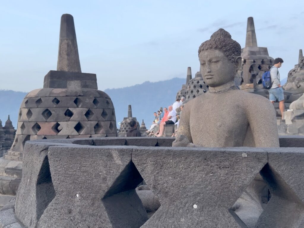 Stone Buddha beside a latticed stupa with mountains on the horizon at Borobudur
