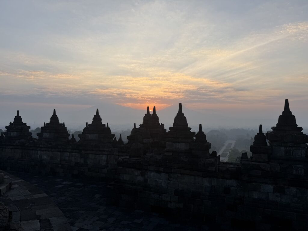 The sun rising behind rows of bell-shaped stupas on the upper terraces of Borobudur, with mist in the valley