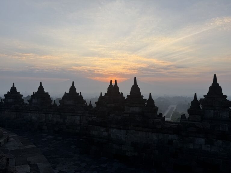 The sun rising behind rows of bell-shaped stupas on the upper terraces of Borobudur, with mist in the valley