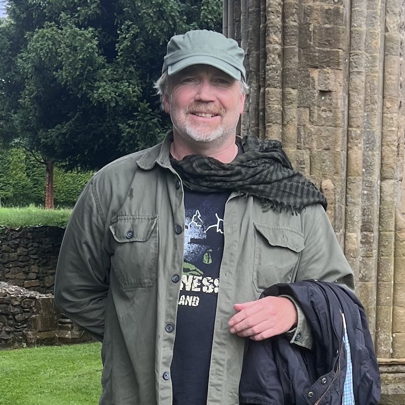 Hugh Newman of Megalithomania in a green cap in front of an old stone wall