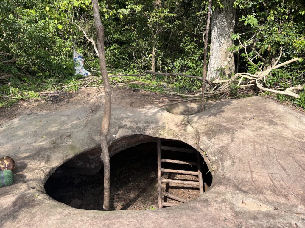An underground shelter under a stone roof with a round skylight opening and a ladder at Phnom Kulen