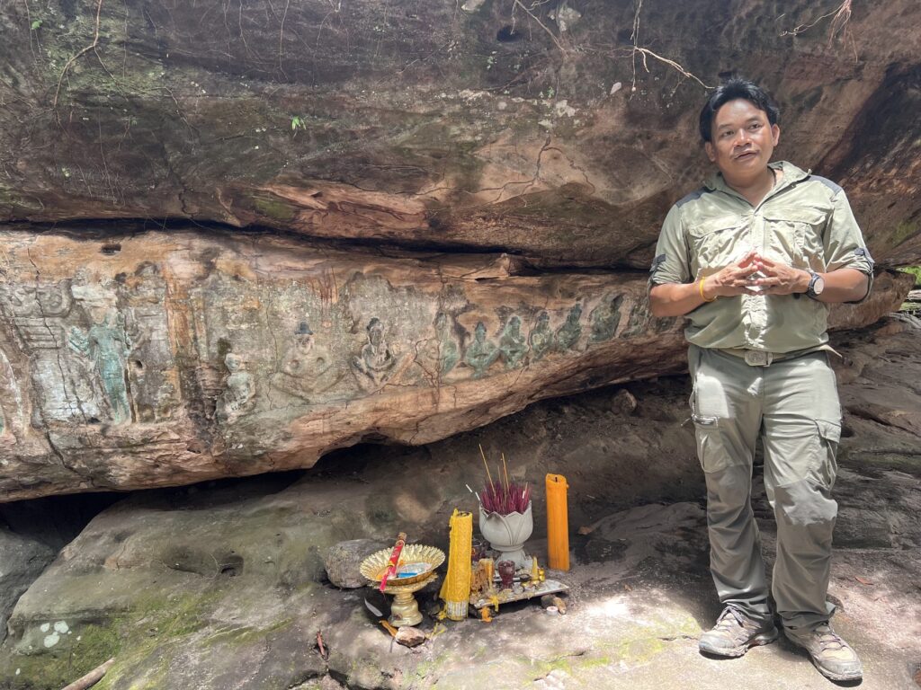 The camp leader standing at a small shrine under a carved rock overhang at Peung Tbal