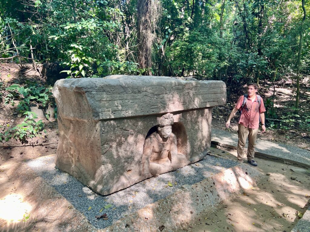 Luke Caverns standing beside a carved Olmec altar with a seated figure in its niche, among the trees at La Venta Park in Villahermosa
