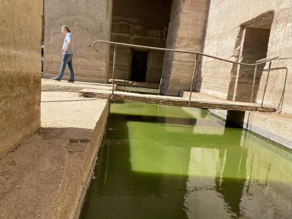 A visitor walks the narrow ledge past a metal footbridge over the green water in the Osirion