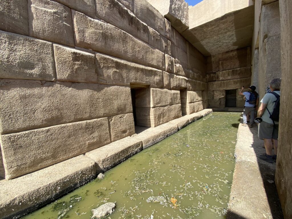 Row of side chamber doorways along a narrow stone ledge and green water channel inside the Osirion